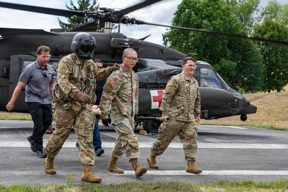 Military personnel in U.S. Army camouflage uniforms are walking away from a UH-60 Black Hawk helicopter marked with a red cross, indicating a medical evacuation (MEDEVAC) configuration. One soldier is wearing a flight helmet and appears to be guiding or escorting another soldier, while two civilians in casual clothing are also present. The scene appears to be a training exercise or operational MEDEVAC scenario on a paved landing area.