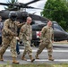 Military personnel in U.S. Army camouflage uniforms are walking away from a UH-60 Black Hawk helicopter marked with a red cross, indicating a medical evacuation (MEDEVAC) configuration. One soldier is wearing a flight helmet and appears to be guiding or escorting another soldier, while two civilians in casual clothing are also present. The scene appears to be a training exercise or operational MEDEVAC scenario on a paved landing area.