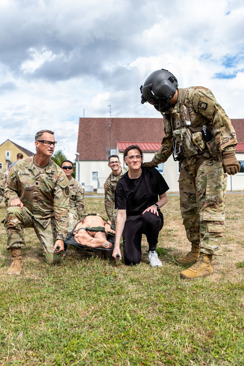 U.S. Army personnel are conducting a medical evacuation or casualty handling training exercise outdoors on a grassy area. A group of soldiers in OCP camouflage uniforms, including one wearing a flight helmet, are gathered around a litter (stretcher) carrying a training mannequin. A civilian or non-uniformed participant in black clothing is also involved in the exercise.
