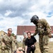 U.S. Army personnel are conducting a medical evacuation or casualty handling training exercise outdoors on a grassy area. A group of soldiers in OCP camouflage uniforms, including one wearing a flight helmet, are gathered around a litter (stretcher) carrying a training mannequin. A civilian or non-uniformed participant in black clothing is also involved in the exercise.