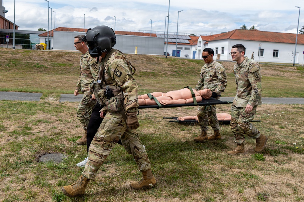 A group of military personnel in US Army MultiCam (OCP) uniforms are conducting a medical evacuation training exercise outdoors on a military installation. Two soldiers are carrying a medical training mannequin on a litter/stretcher, while two others walk alongside. The setting appears to be a European military base, identifiable by the architecture with red-tiled roofs in the background.
