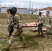 A group of military personnel in US Army MultiCam (OCP) uniforms are conducting a medical evacuation training exercise outdoors on a military installation. Two soldiers are carrying a medical training mannequin on a litter/stretcher, while two others walk alongside. The setting appears to be a European military base, identifiable by the architecture with red-tiled roofs in the background.
