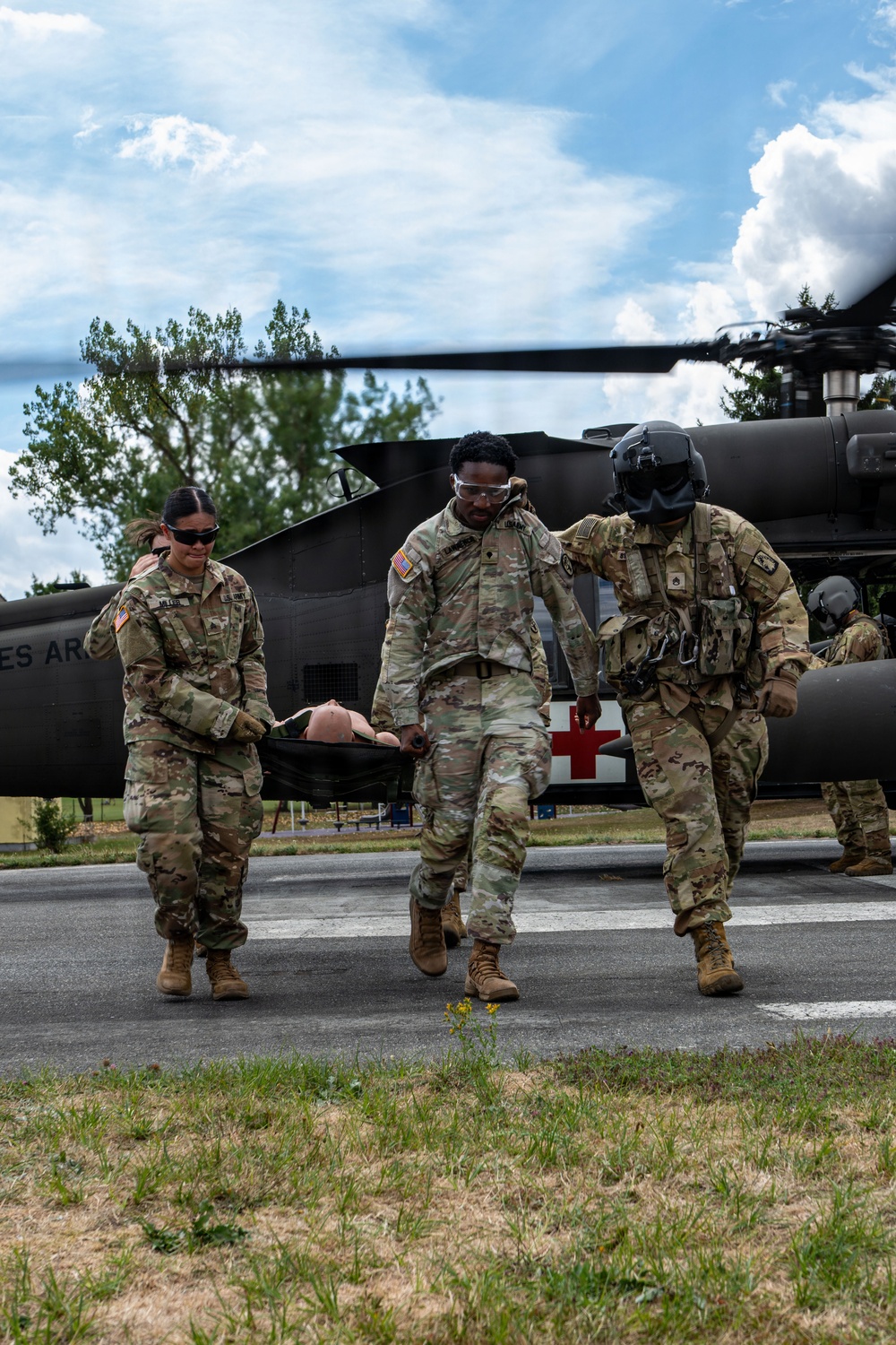 Three U.S. Army soldiers in OCP (Operational Camouflage Pattern) uniforms are carrying a litter (stretcher) with what appears to be a training mannequin away from a military medevac helicopter marked with a red cross. The helicopter in the background is a UH-60 Black Hawk configured for medical evacuation, identifiable by the red cross insignia on its fuselage. This appears to be a medical evacuation training exercise conducted on a paved landing zone.