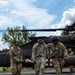 Three U.S. Army soldiers in OCP (Operational Camouflage Pattern) uniforms are carrying a litter (stretcher) with what appears to be a training mannequin away from a military medevac helicopter marked with a red cross. The helicopter in the background is a UH-60 Black Hawk configured for medical evacuation, identifiable by the red cross insignia on its fuselage. This appears to be a medical evacuation training exercise conducted on a paved landing zone.