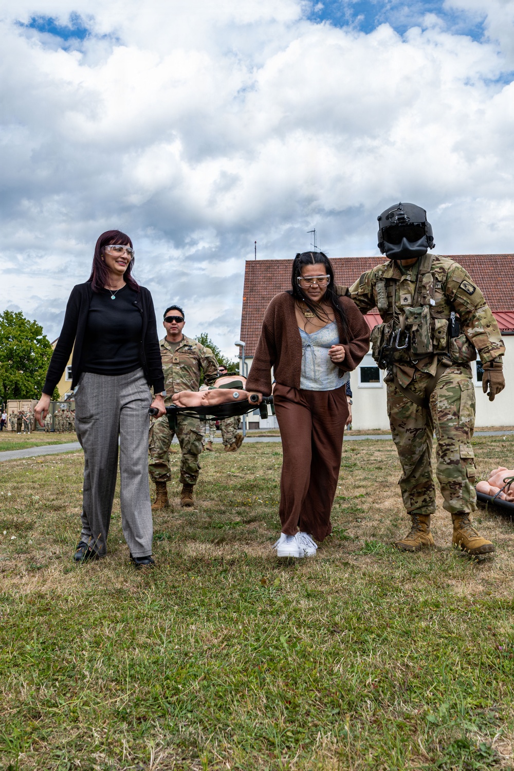 A military training exercise or demonstration is taking place on a grassy field near a building with a red tile roof. Two civilian women wearing safety glasses are participating alongside uniformed soldiers in carrying a stretcher with a training mannequin. The scene appears to be a familiarization or community engagement event where civilians are invited to experience military medical evacuation procedures.