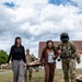 A military training exercise or demonstration is taking place on a grassy field near a building with a red tile roof. Two civilian women wearing safety glasses are participating alongside uniformed soldiers in carrying a stretcher with a training mannequin. The scene appears to be a familiarization or community engagement event where civilians are invited to experience military medical evacuation procedures.