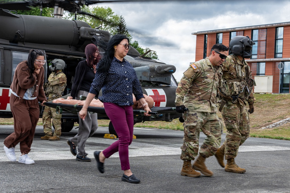A group of military personnel and civilians are carrying a stretcher with a simulated casualty (mannequin) away from a military medevac helicopter marked with a Red Cross symbol. The scene appears to be a medical evacuation training exercise conducted on a paved area near a building. Both uniformed soldiers in camouflage and civilians in casual clothing are participating in the drill.
