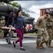 A group of military personnel and civilians are carrying a stretcher with a simulated casualty (mannequin) away from a military medevac helicopter marked with a Red Cross symbol. The scene appears to be a medical evacuation training exercise conducted on a paved area near a building. Both uniformed soldiers in camouflage and civilians in casual clothing are participating in the drill.