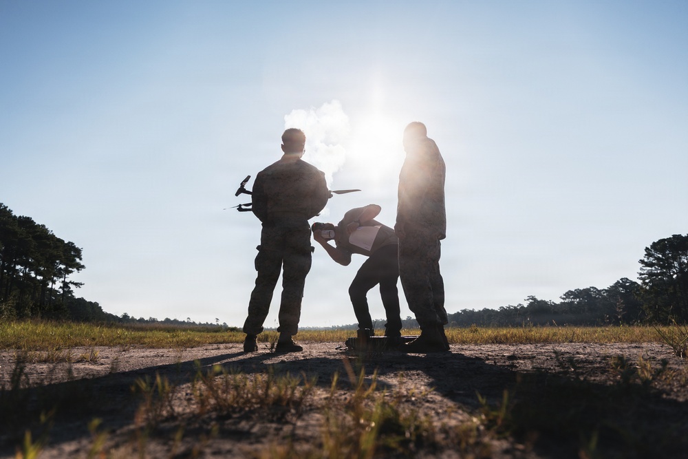 Three individuals in military-style camouflage uniforms are gathered in an open field, appearing to prepare or inspect a small drone/UAV. The image is shot against a bright backlit sun, creating strong silhouettes of the figures. The setting appears to be a rural or training area with treelines visible in the background.