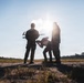 Three individuals in military-style camouflage uniforms are gathered in an open field, appearing to prepare or inspect a small drone/UAV. The image is shot against a bright backlit sun, creating strong silhouettes of the figures. The setting appears to be a rural or training area with treelines visible in the background.