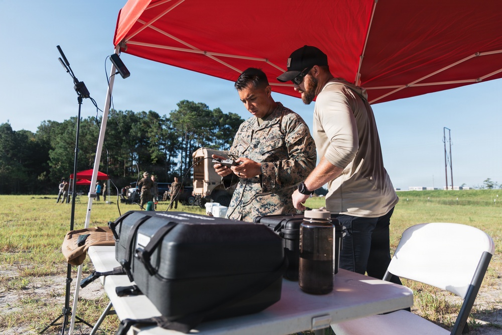 Two individuals, one in U.S. Marine Corps digital camouflage uniform and one in civilian tactical attire, are working together under a red pop-up canopy tent in an open field. They appear to be operating or configuring a handheld drone controller or electronic device at a folding table. The setting suggests a military field exercise or drone operations training event.