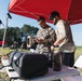 Two individuals, one in U.S. Marine Corps digital camouflage uniform and one in civilian tactical attire, are working together under a red pop-up canopy tent in an open field. They appear to be operating or configuring a handheld drone controller or electronic device at a folding table. The setting suggests a military field exercise or drone operations training event.