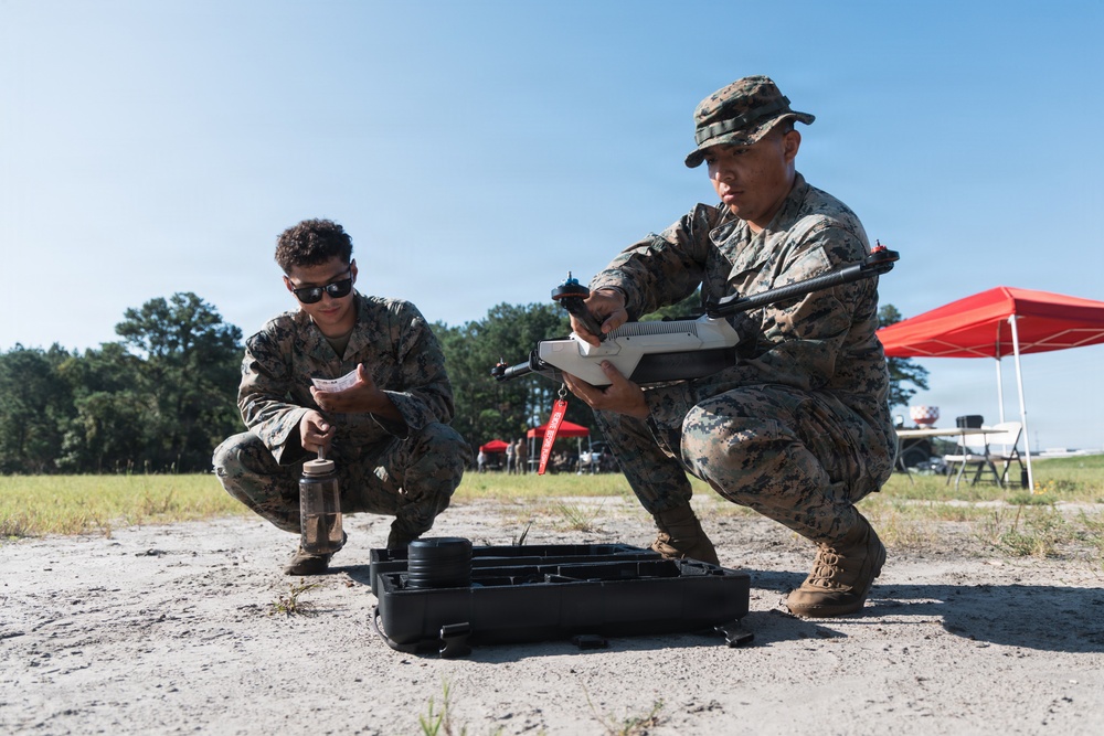 Two military personnel in MARPAT (Marine digital camouflage) uniforms are crouched on sandy ground preparing what appears to be a small unmanned aerial vehicle (drone). One service member holds the drone while the other reviews a document or checklist. The setting appears to be an outdoor field exercise or demonstration area with a red canopy tent visible in the background.