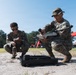 Two military personnel in MARPAT (Marine digital camouflage) uniforms are crouched on sandy ground preparing what appears to be a small unmanned aerial vehicle (drone). One service member holds the drone while the other reviews a document or checklist. The setting appears to be an outdoor field exercise or demonstration area with a red canopy tent visible in the background.
