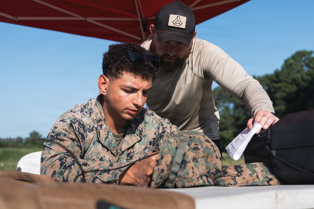 A U.S. Marine in MARPAT digital camouflage uniform is seated at a table outdoors, appearing to review or work on materials. A civilian or contractor in tan clothing and a black cap is leaning over, pointing at or handing a document to the Marine. The setting appears to be an outdoor training or administrative station under a red canopy.