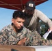 A U.S. Marine in MARPAT digital camouflage uniform is seated at a table outdoors, appearing to review or work on materials. A civilian or contractor in tan clothing and a black cap is leaning over, pointing at or handing a document to the Marine. The setting appears to be an outdoor training or administrative station under a red canopy.