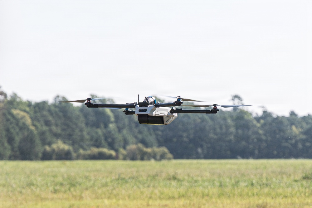 A quadcopter drone is captured mid-flight over an open grassy field with a treeline visible in the background. The drone features a white central body/payload module mounted on a black carbon fiber frame with four rotors. The image is sharp and well-exposed, with the drone in clear focus against a softly blurred background.