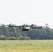 A quadcopter drone is captured mid-flight over an open grassy field with a treeline visible in the background. The drone features a white central body/payload module mounted on a black carbon fiber frame with four rotors. The image is sharp and well-exposed, with the drone in clear focus against a softly blurred background.