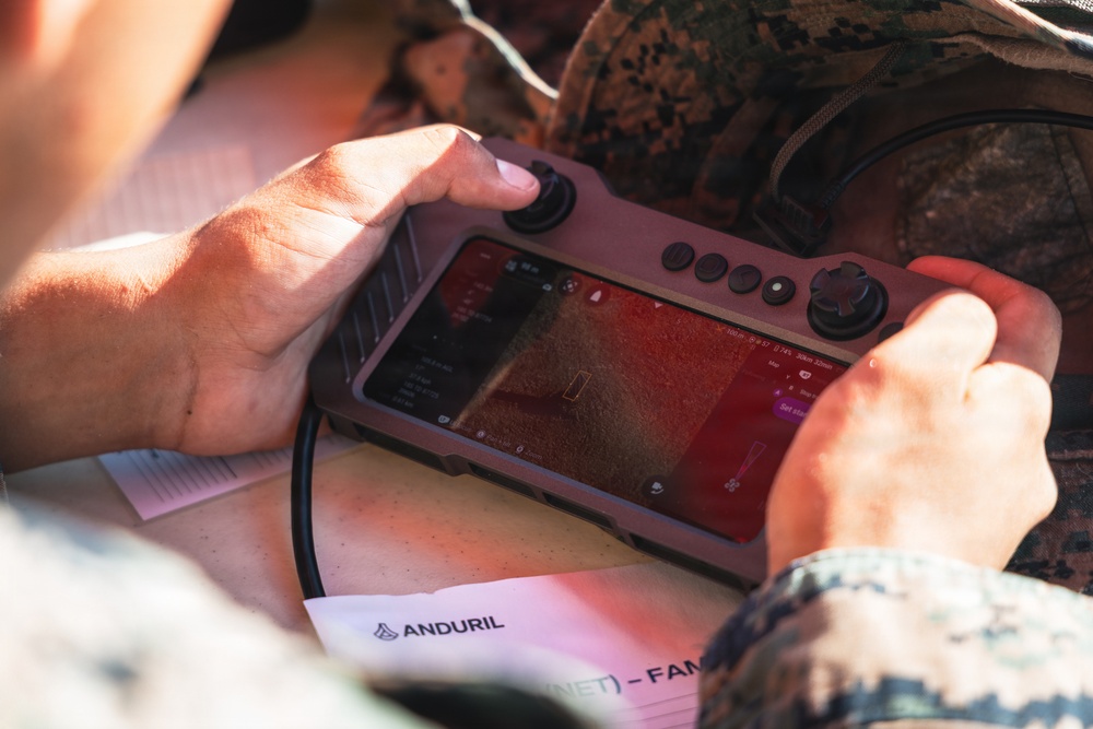 A person in military camouflage uniform is holding a handheld drone or autonomous system controller manufactured by Anduril Industries. The device features a touchscreen display showing what appears to be a camera feed or targeting interface, flanked by joysticks and control buttons. A document bearing the Anduril logo is visible beneath the controller.