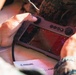 A person in military camouflage uniform is holding a handheld drone or autonomous system controller manufactured by Anduril Industries. The device features a touchscreen display showing what appears to be a camera feed or targeting interface, flanked by joysticks and control buttons. A document bearing the Anduril logo is visible beneath the controller.