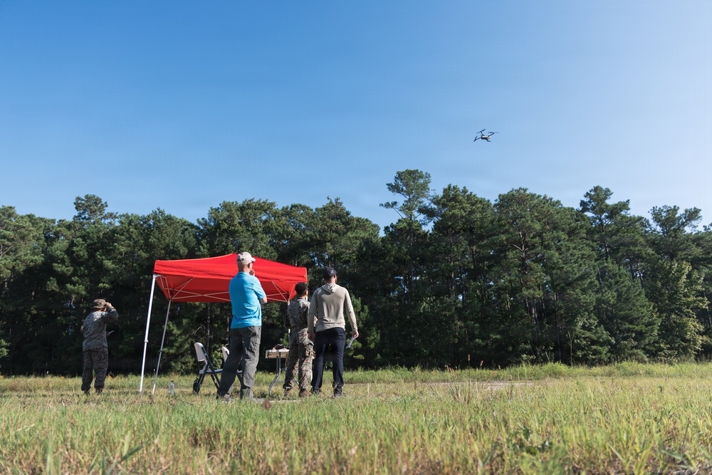 A group of four individuals are gathered in an open grassy field operating a small drone, which is visible in flight against a clear blue sky. A red pop-up canopy tent is set up nearby with a folding table and chair, suggesting an organized outdoor operation or demonstration. Two of the individuals appear to be wearing military camouflage uniforms, while the others are in civilian clothing.