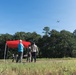 A group of four individuals are gathered in an open grassy field operating a small drone, which is visible in flight against a clear blue sky. A red pop-up canopy tent is set up nearby with a folding table and chair, suggesting an organized outdoor operation or demonstration. Two of the individuals appear to be wearing military camouflage uniforms, while the others are in civilian clothing.