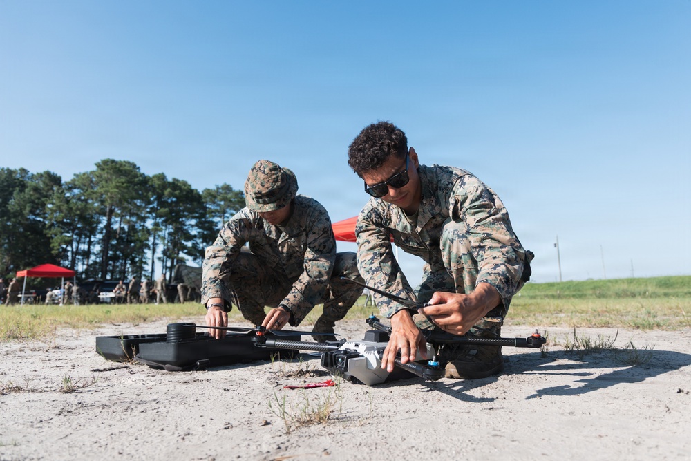 Two military personnel in MARPAT (Marine pattern) camouflage uniforms are crouching on sandy ground, assembling or preparing a multi-rotor drone/UAV for operation. A black equipment case is visible nearby, suggesting the drone was recently unpacked. In the background, additional personnel and a red canopy tent are visible at what appears to be a field training or demonstration site.