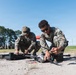 Two military personnel in MARPAT (Marine pattern) camouflage uniforms are crouching on sandy ground, assembling or preparing a multi-rotor drone/UAV for operation. A black equipment case is visible nearby, suggesting the drone was recently unpacked. In the background, additional personnel and a red canopy tent are visible at what appears to be a field training or demonstration site.