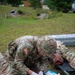 A U.S. Army soldier in OCP (Operational Camouflage Pattern) uniform is performing tactical combat casualty care (TCCC) on a simulated casualty lying on the ground outdoors. The medic is wearing latex gloves and working with medical supplies laid out on a blue pad. A second simulated casualty with mock wounds is visible on a litter in the background, suggesting a field medical training exercise.