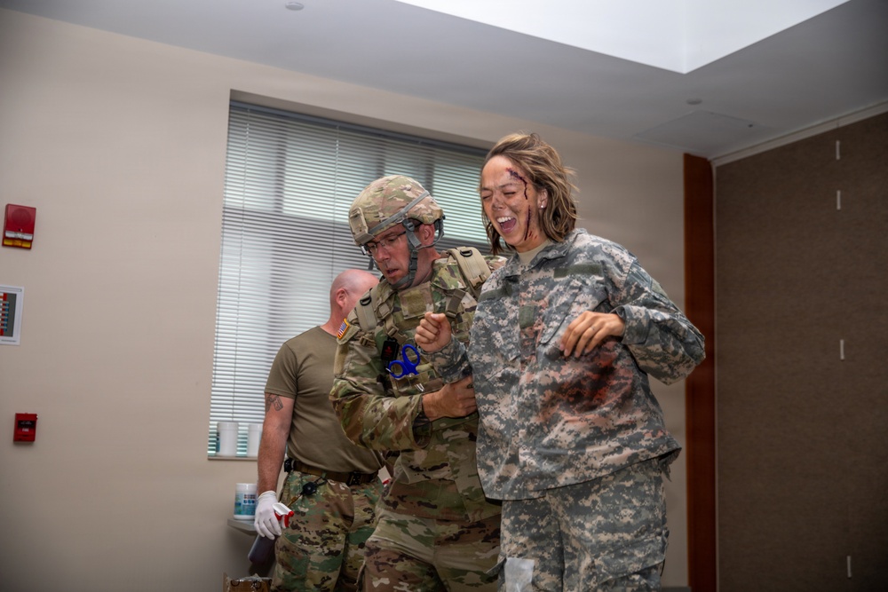 The image depicts what appears to be a military medical training exercise or simulation. A soldier in full combat gear and helmet is assisting a female soldier wearing simulated injury makeup (moulage) on her face and uniform. A third individual in a tan t-shirt is visible in the background observing the scenario.