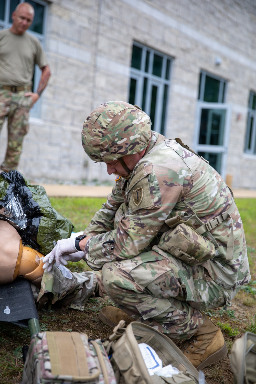 A U.S. Army soldier in OCP (Operational Camouflage Pattern) uniform and helmet is kneeling on grass performing tactical combat casualty care (TCCC) on a training mannequin. The soldier is wearing white medical gloves and appears to be applying a tourniquet or bandage to the mannequin's limb. A second soldier in a t-shirt observes in the background, suggesting this is a training exercise.