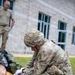 A U.S. Army soldier in OCP (Operational Camouflage Pattern) uniform and helmet is kneeling on grass performing tactical combat casualty care (TCCC) on a training mannequin. The soldier is wearing white medical gloves and appears to be applying a tourniquet or bandage to the mannequin's limb. A second soldier in a t-shirt observes in the background, suggesting this is a training exercise.