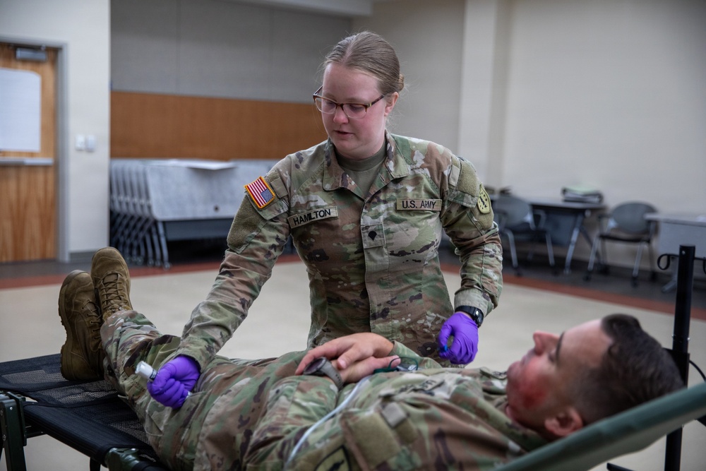 A U.S. Army soldier named Hamilton is performing medical treatment or assessment on another soldier lying on a field stretcher. The standing soldier is wearing purple medical gloves and appears to be conducting a tactical combat casualty care (TCCC) exercise or training scenario. The setting appears to be an indoor training facility.