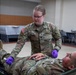 A U.S. Army soldier named Hamilton is performing medical treatment or assessment on another soldier lying on a field stretcher. The standing soldier is wearing purple medical gloves and appears to be conducting a tactical combat casualty care (TCCC) exercise or training scenario. The setting appears to be an indoor training facility.