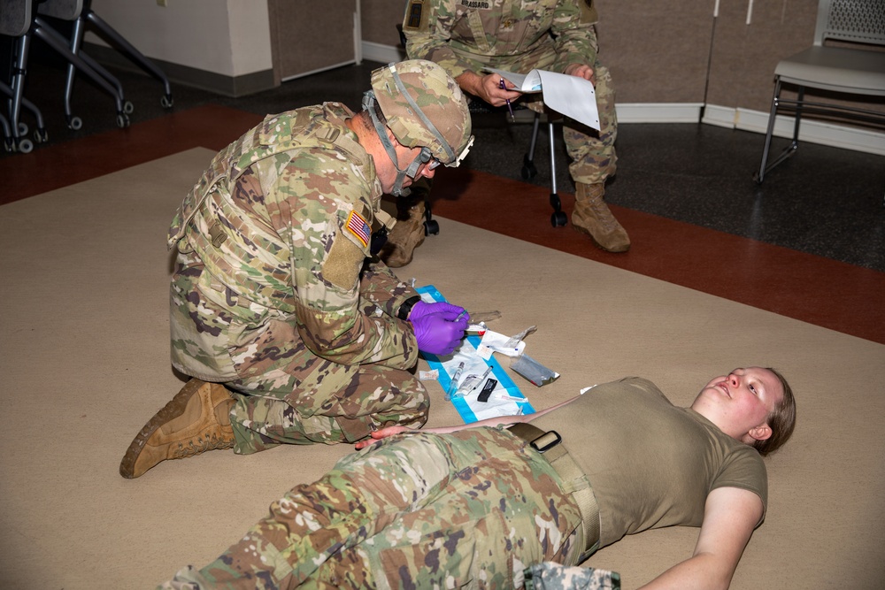 A U.S. Army soldier wearing OCP (Operational Camouflage Pattern) uniform and purple medical gloves is performing a medical training exercise on another soldier lying on the floor. A third soldier in the background appears to be evaluating or grading the exercise while holding a clipboard and pen. The setting appears to be an indoor training facility or classroom.
