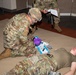 A U.S. Army soldier wearing OCP (Operational Camouflage Pattern) uniform and purple medical gloves is performing a medical training exercise on another soldier lying on the floor. A third soldier in the background appears to be evaluating or grading the exercise while holding a clipboard and pen. The setting appears to be an indoor training facility or classroom.