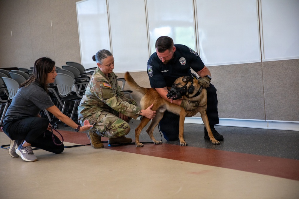 Three individuals are interacting with a muzzled Belgian Malinois or German Shepherd-type dog in what appears to be an indoor meeting or training room. The group includes a civilian woman, a military service member in camouflage uniform, and a law enforcement officer in a dark uniform. The setting suggests a K-9 demonstration, training session, or community outreach event.