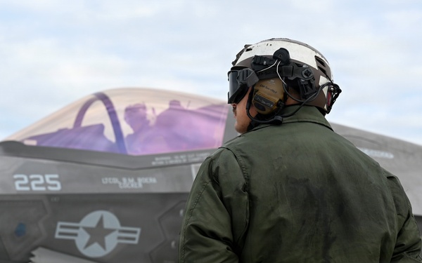 A military ground crew member in a green flight suit and helmet stands on a tarmac with hands clasped behind their back, observing an F-35 fighter jet marked with the number '225' and 'VMFA' designation, indicating a Marine Fighter Attack Squadron. The aircraft displays a US military star-and-bar insignia and has its canopy open with a pilot visible inside the cockpit. The scene appears to be a pre-flight or post-flight inspection on a military airfield.