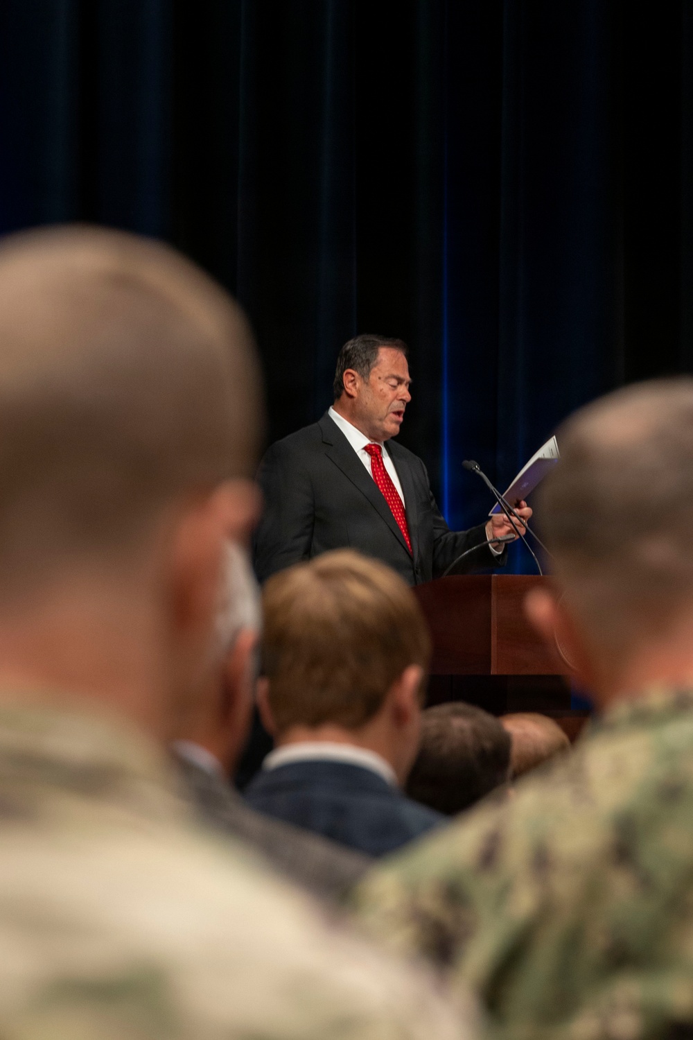A man in a dark suit with a red tie is speaking at a wooden podium, holding what appears to be a document or booklet. He is addressing an audience that includes individuals in military uniforms, visible in the foreground. The setting appears to be a formal ceremony or official briefing in a venue with dark blue curtain backdrop.