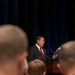 A man in a dark suit with a red tie is speaking at a wooden podium, holding what appears to be a document or booklet. He is addressing an audience that includes individuals in military uniforms, visible in the foreground. The setting appears to be a formal ceremony or official briefing in a venue with dark blue curtain backdrop.