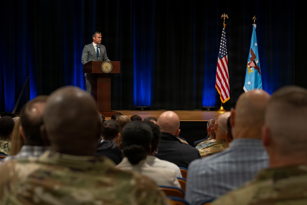 A civilian male in a gray suit is delivering a speech at a wooden podium bearing an official seal, addressing a mixed audience of military personnel and civilians in a formal auditorium setting. The stage is flanked by the United States flag and what appears to be a Department of Defense or military branch flag on a blue light-illuminated backdrop. The audience includes individuals in military camouflage uniforms as well as civilian attire.