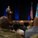 A civilian male in a gray suit is delivering a speech at a wooden podium bearing an official seal, addressing a mixed audience of military personnel and civilians in a formal auditorium setting. The stage is flanked by the United States flag and what appears to be a Department of Defense or military branch flag on a blue light-illuminated backdrop. The audience includes individuals in military camouflage uniforms as well as civilian attire.