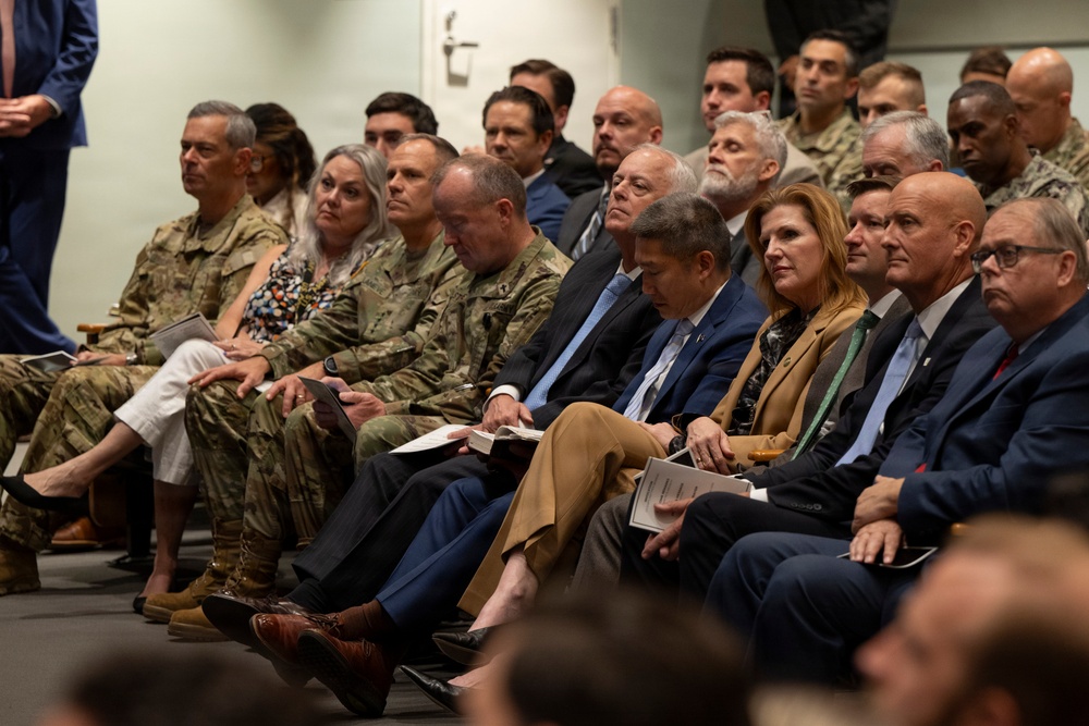 A formal gathering featuring a mixed audience of military personnel in camouflage uniforms and civilians in business attire, seated in rows as attendees of what appears to be a briefing, ceremony, or official presentation. The front rows include senior military officers alongside suited civilians, suggesting a high-level official event. Several attendees are holding printed documents or programs.
