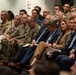 A formal gathering featuring a mixed audience of military personnel in camouflage uniforms and civilians in business attire, seated in rows as attendees of what appears to be a briefing, ceremony, or official presentation. The front rows include senior military officers alongside suited civilians, suggesting a high-level official event. Several attendees are holding printed documents or programs.