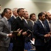 A group of formally dressed individuals, predominantly men in business suits, are standing in rows and appear to be participating in a religious or memorial service. One individual in the center is wearing a dark military or naval uniform with gold sleeve stripes. Most attendees are holding books or printed programs and appear to be singing or reciting something in unison.