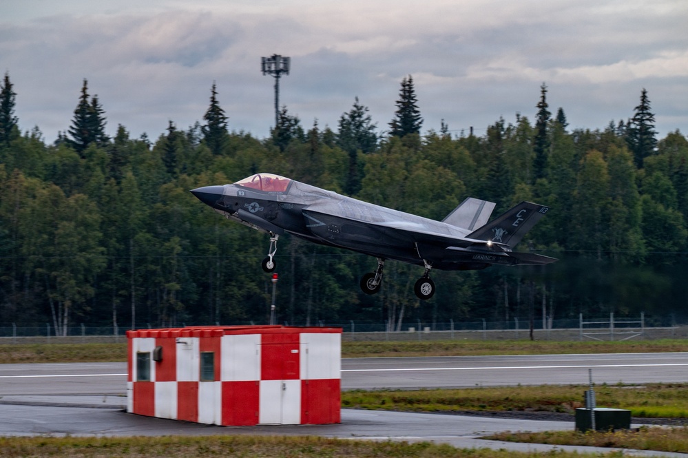 A U.S. Marines F-35B Lightning II stealth fighter jet is captured during takeoff or low-altitude flight over a runway. The aircraft displays Marine Corps markings and tail codes, with landing gear still deployed. The setting appears to be a northern airfield surrounded by dense coniferous forest under overcast skies.