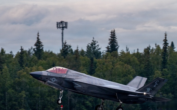 A U.S. Marines F-35B Lightning II stealth fighter jet is captured during takeoff or low-altitude flight over a runway. The aircraft displays Marine Corps markings and tail codes, with landing gear still deployed. The setting appears to be a northern airfield surrounded by dense coniferous forest under overcast skies.