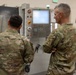 Two military personnel in MultiCam/OCP camouflage uniforms are standing in front of a Haas CNC machine in what appears to be a machining or fabrication facility. The individual on the right, who has gray hair and appears to be of higher rank based on visible shoulder insignia, is observing with arms crossed while the other person faces the machine's control panel. They appear to be engaged in a briefing or inspection of the equipment.