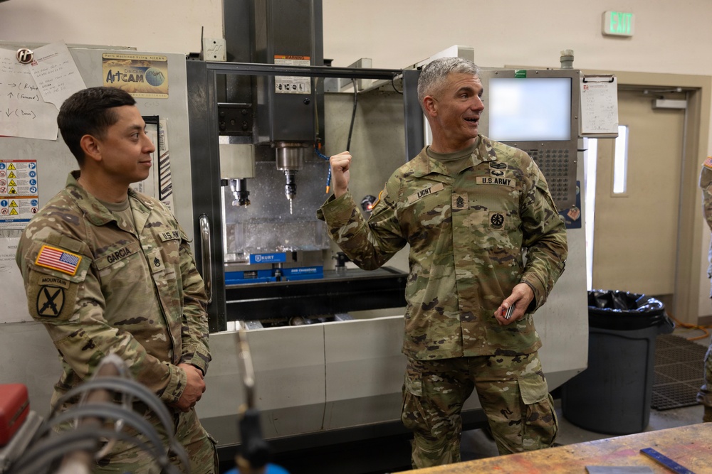 Two U.S. Army soldiers in OCP (Operational Camouflage Pattern) uniforms are standing in what appears to be a machining or fabrication facility. The soldier on the right, identified by name tape as 'LIGHT,' appears to be speaking or explaining something, while the soldier on the left, identified as 'GARCIA,' listens. A CNC milling machine is visible in the background.