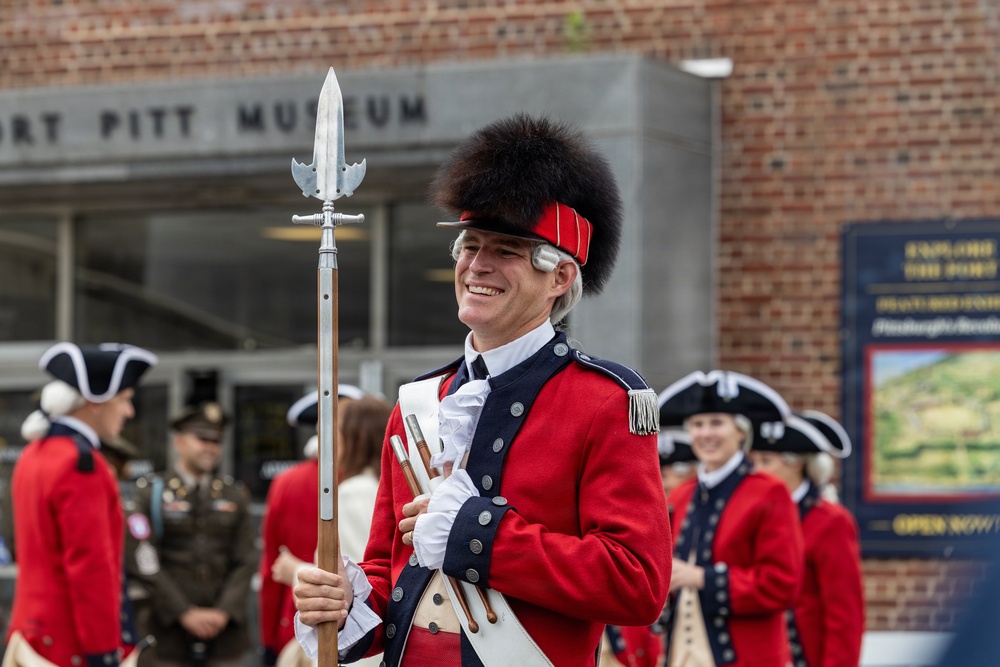 A group of historical reenactors dressed in 18th-century British or colonial military uniforms are gathered outside the Fort Pitt Museum. The central figure, smiling broadly, wears a red coat with blue facings, a tall bearskin cap, white gloves, and carries a halberd or spontoon. The scene appears to be a ceremonial or commemorative event at the museum.